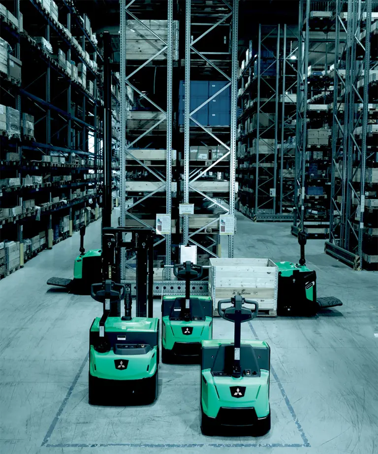 Flera gröna Mitsubishi eldrivna plocktruckar uppställda framför höga pallställ i lager med blåtonad belysning – Logisnext