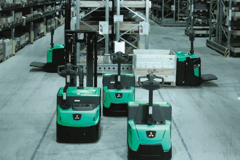 Flera gröna Mitsubishi eldrivna plocktruckar uppställda framför höga pallställ i lager – Logisnext