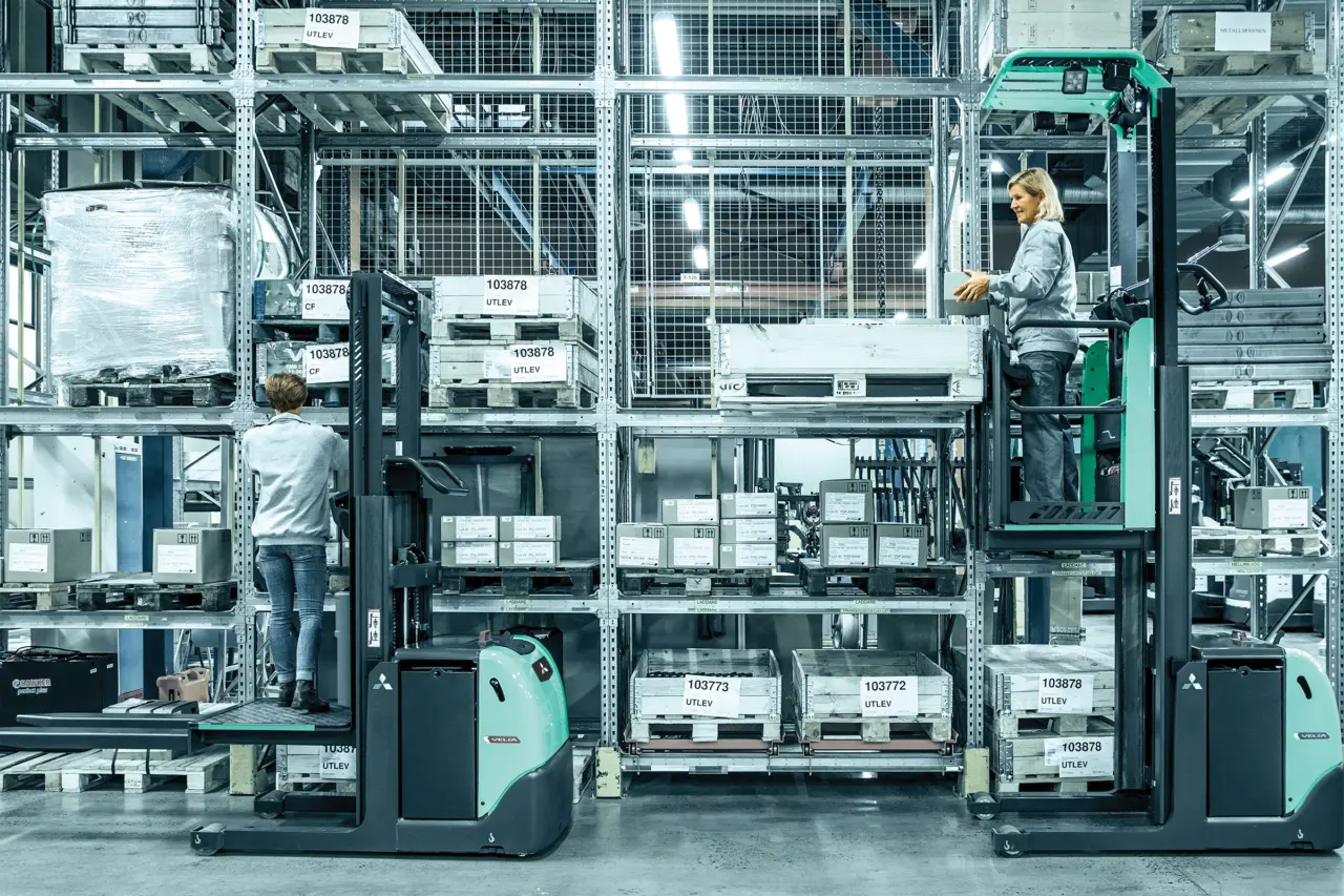 Två lagerarbetare som plockar gods på olika höjder med var sin Logisnext VELIA orderplocktruck i ett flervånings lagersystem