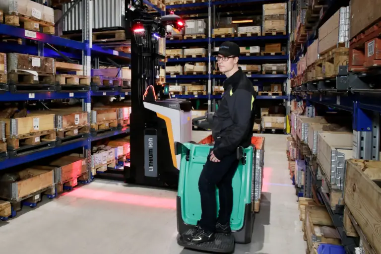 Logisnext-medarbetare som åker på en autonom Rocla reachtruck med Li-ion-batteri i en smal lagergång med rött säkerhetsljus på golvet