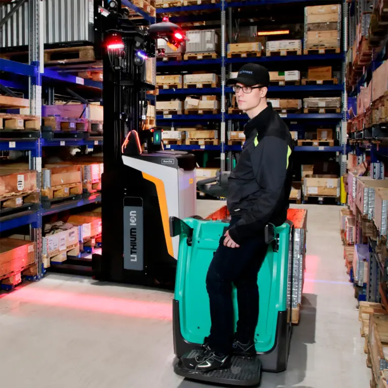 Man med Logisnext-keps kör ståplattform-truck förbi Rocla autonom litiumtruck med röda varningsljus i pallställslager – Logisnext