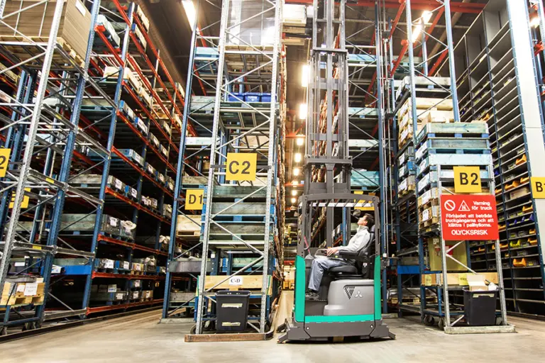 Trucksförare i en Mitsubishi reachtruck vid höga pallställ märkta med gångnummer i ett lager