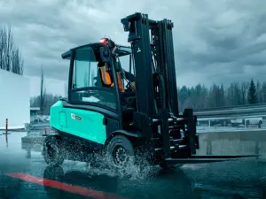 Logisnext EDiA eltruck på 5 ton med sluten hytt och gaffelpositionerare som kör genom vattenpölar på en våt industrigård under mörka regnmoln