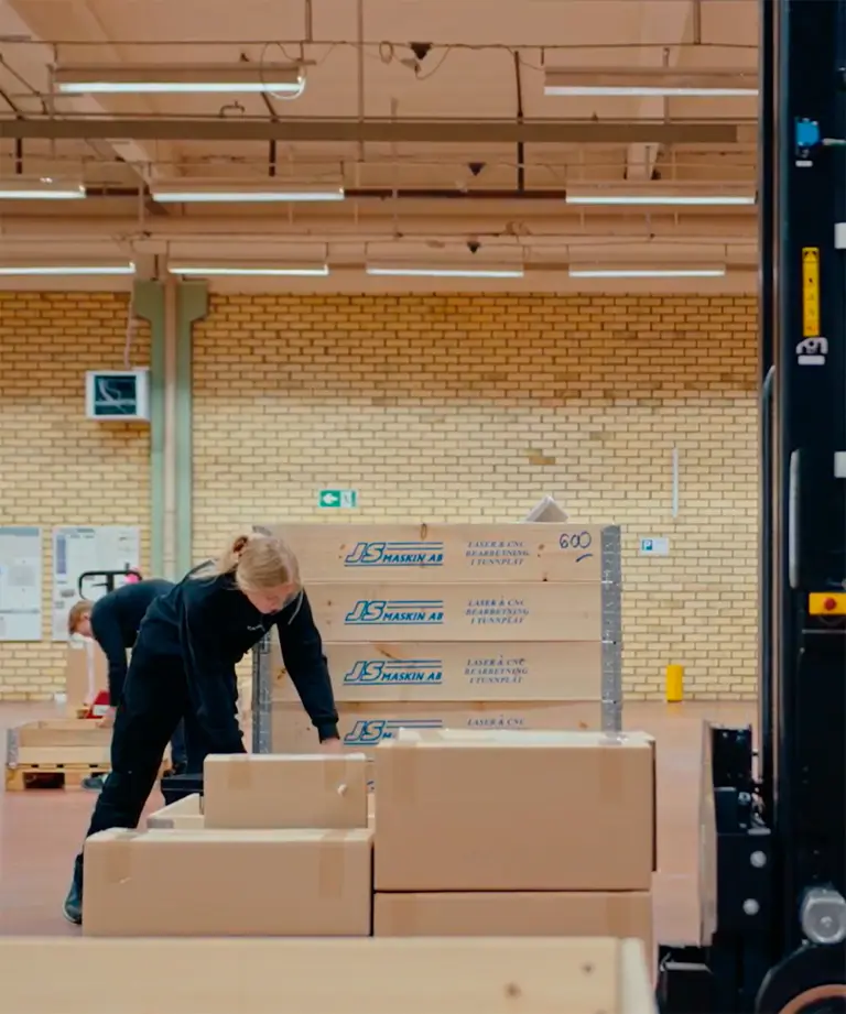Medarbetare som packar kartonger på pallar i en lagerhall, med en truck med lyftmast i förgrunden
