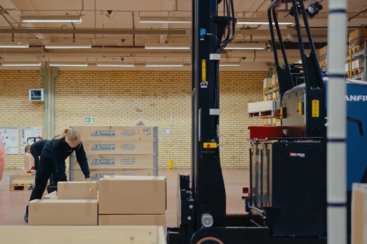 Medarbetare som packar kartonger på pallar i en lagerhall, med en truck med lyftmast i förgrunden