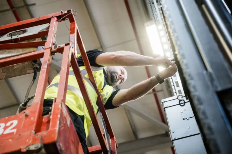 Man i varselväst reparerar hyllställning från en saxlift på ett lager