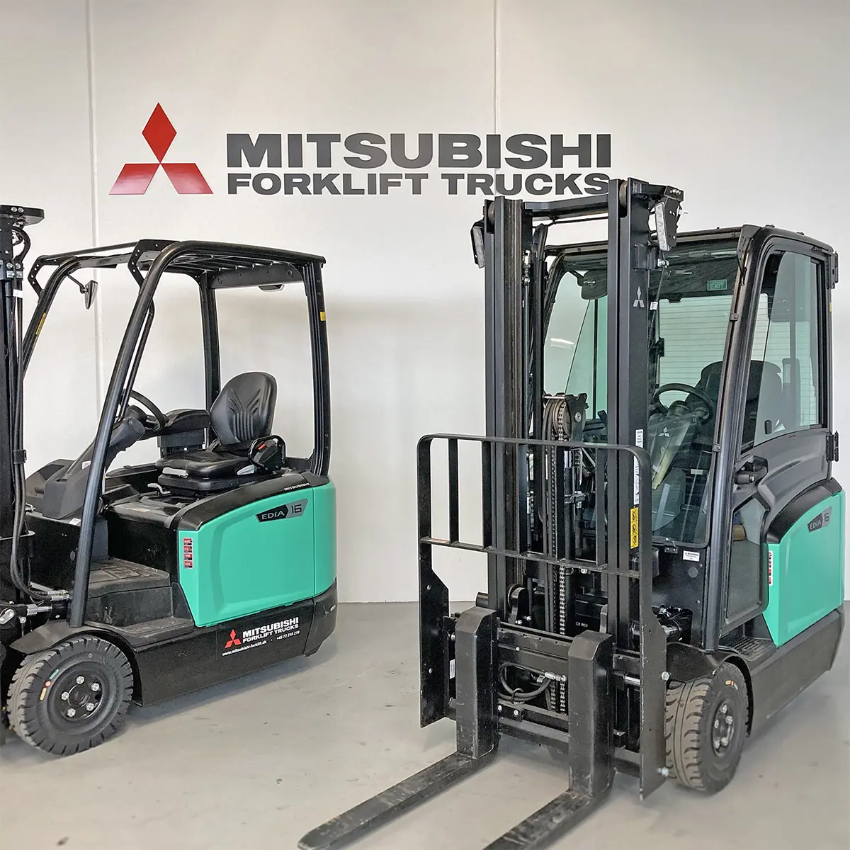 Två Mitsubishi Edia motviktstruckar framför Mitsubishi Forklift Trucks-logotyp i showroom – Logisnext
