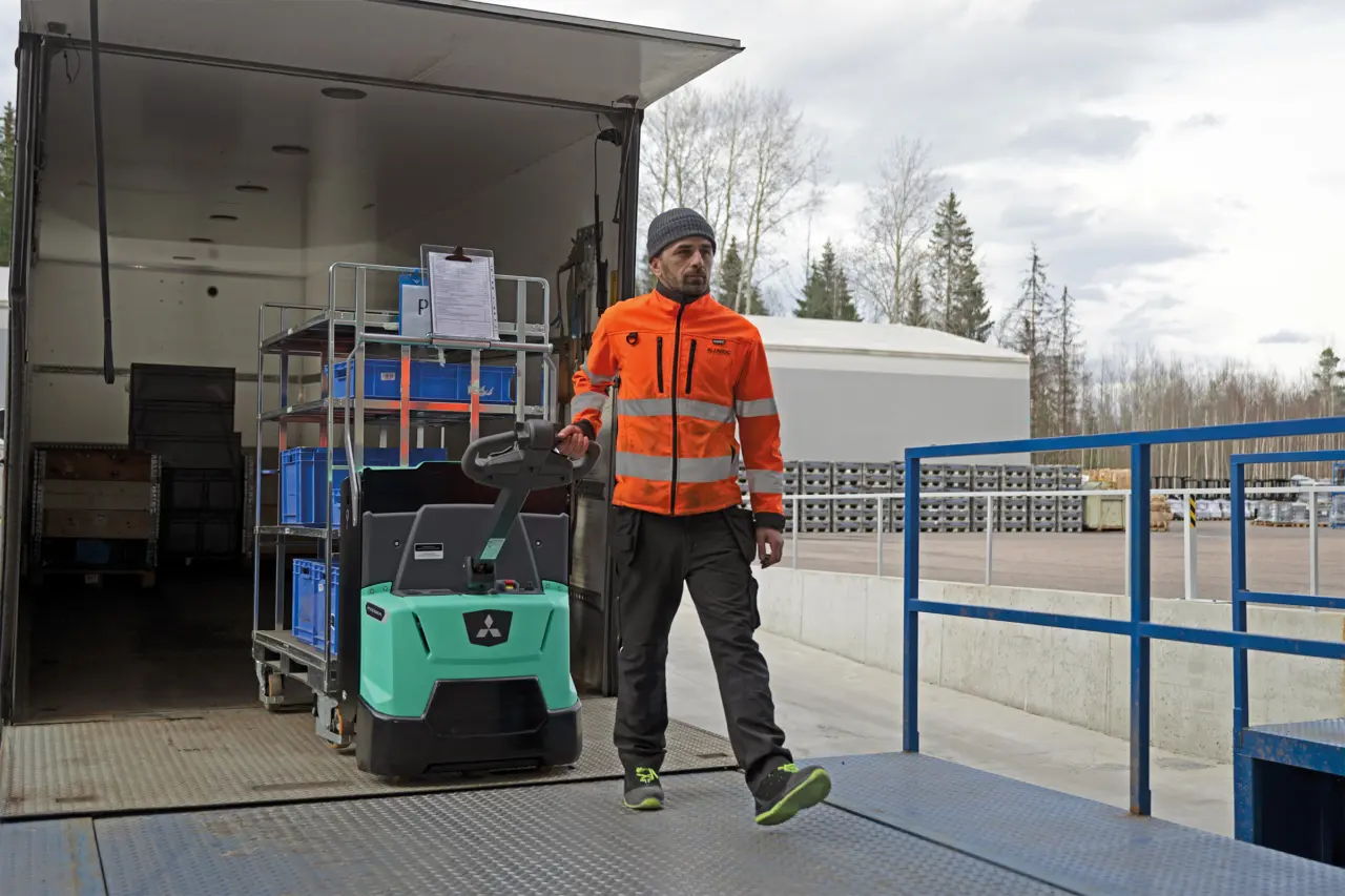 Lagerarbetare i varselkläder som lossar en rullcontainer med blå backar från en lastbil med en gående eldriven pallyftare ur Logisnext Premia-serien