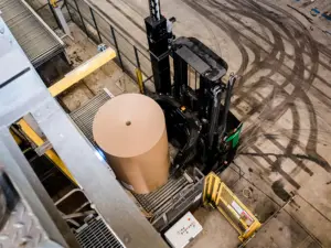Vy ovanifrån av en truck med klämmar som hanterar en stor pappersrulle på ett golv med däckspår